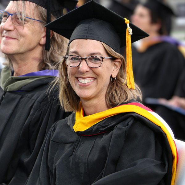 La profesora Angela Redmon, ataviada con la toga de graduación, se encuentra sentada junto a otros miembros del cuerpo docente en una ceremonia de graduación.