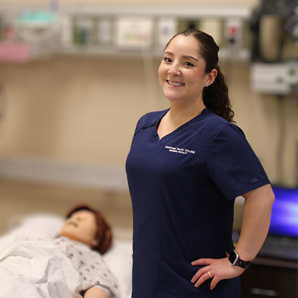 Carolina Martínez Calderón lleva una bata quirúrgica azul oscuro y se encuentra de pie junto a un maniquí en el laboratorio de enfermería.