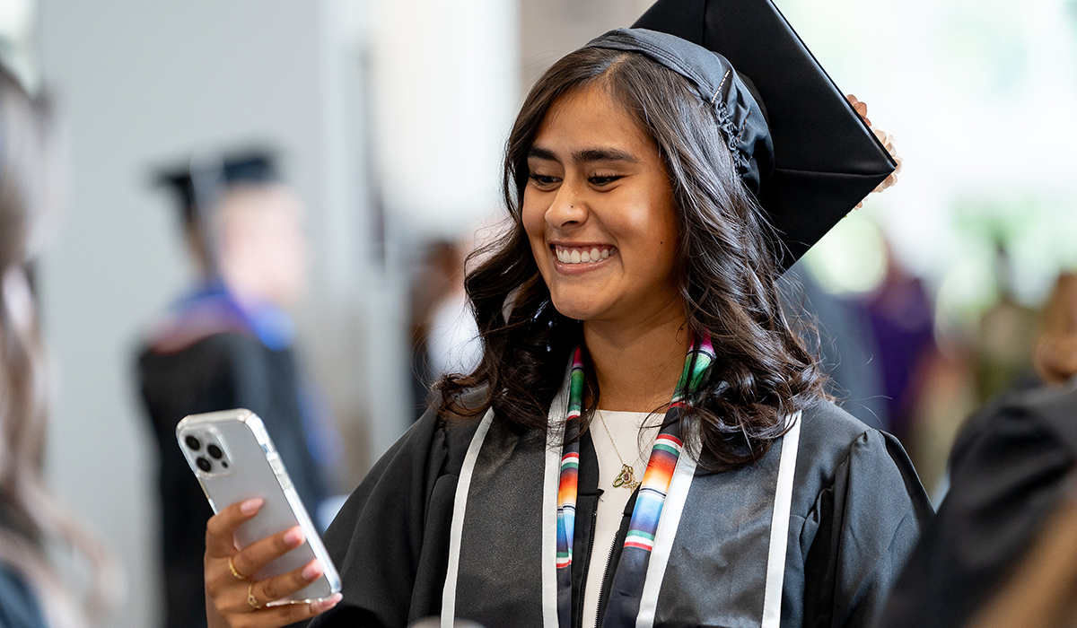 Una graduada, ataviada con toga y birrete, sonríe mientras mira su teléfono.