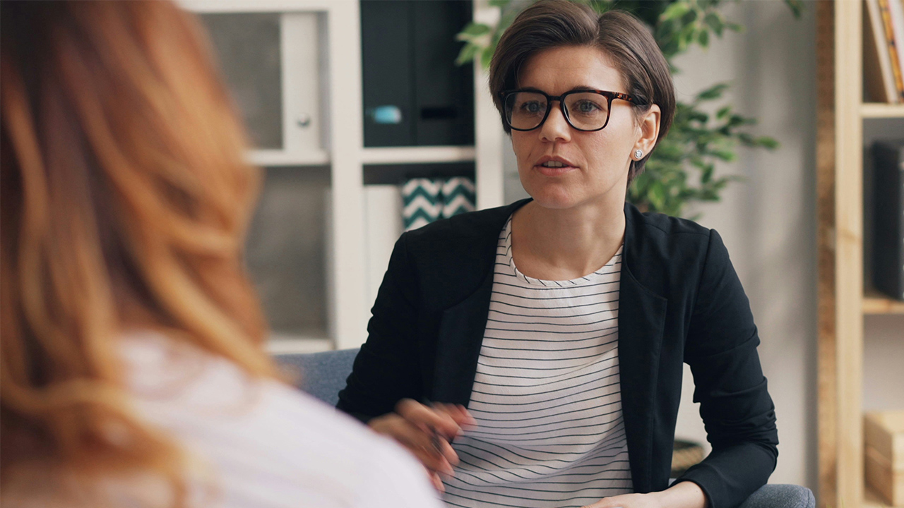 Una terapeuta, sentada en un sofá, habla con una paciente que está sentada frente a ella.
