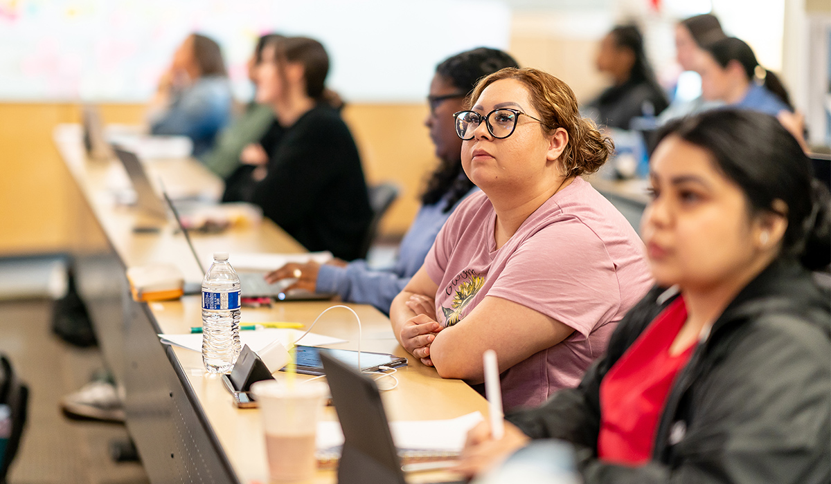 Un grupo de alumnos está sentado en mesas largas y mira hacia la parte delantera del aula.