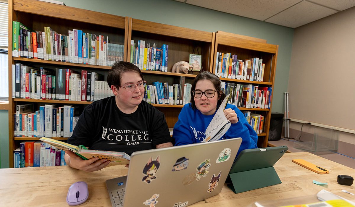Dos estudiantes están sentados a una mesa frente a un ordenador portátil y miran juntos un libro.