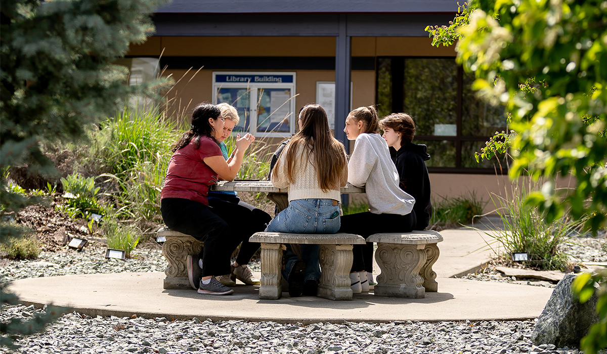 Un grupo de estudiantes está sentado alrededor de una mesa redonda de hormigón en un jardín de plantas autóctonas.