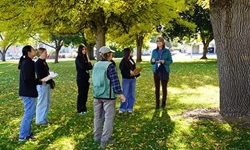 La Dra. Joan Qazi posa bajo un árbol junto a un grupo de estudiantes durante una actividad con motivo del Día del Árbol.