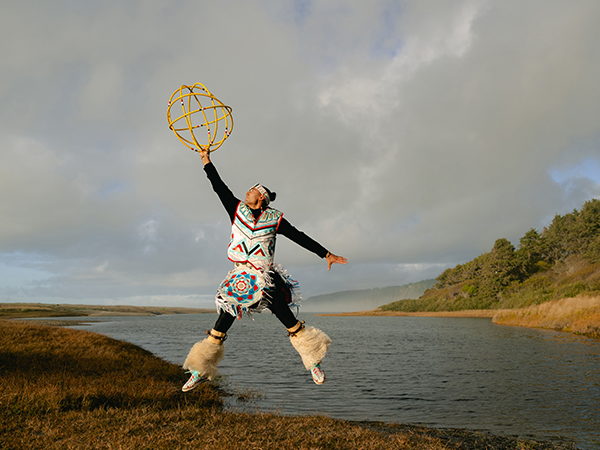 El bailarín de aros Eric Hernández, ataviado con trajes tradicionales, se encuentra junto a un río y salta en el aire sosteniendo varios aros.