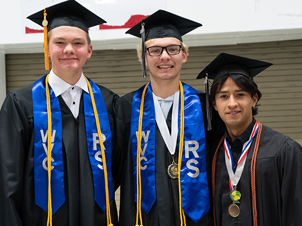 Tres graduados de WVC Omak, vestidos con togas negras, estolas azules y cordones de honor, sonríen a la cámara.
