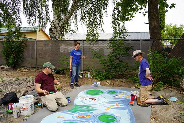 Ellen Bruex se encuentra junto a un mural de un estanque en proceso de realización, acompañada de dos alumnos de la clase de muralismo.