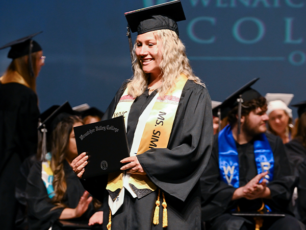 Una graduada de WVC Omak, ataviada con una estola dorada, cruza el escenario sosteniendo su título.