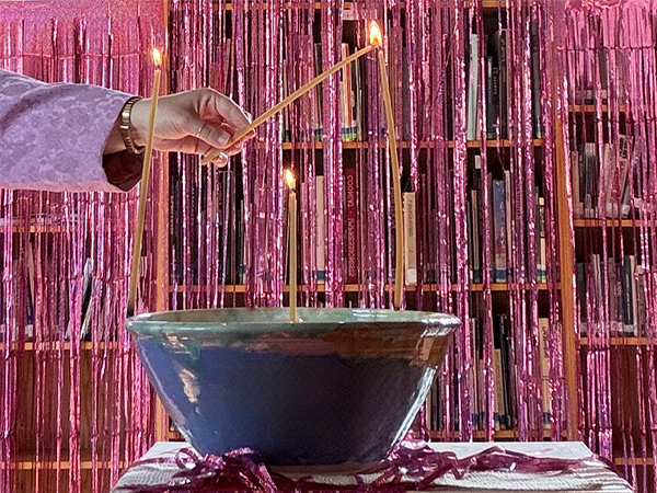 En un cuenco de cerámica azul y verde hay tres velas apagadas, y una mano sostiene una vela encendida junto a la mecha de una vela apagada.