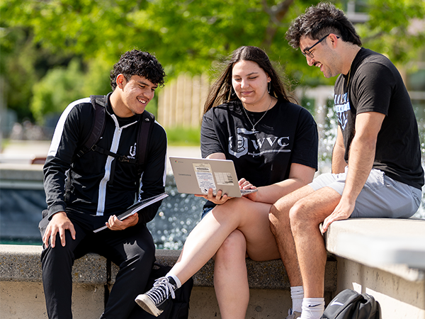 Tres estudiantes de la WVC, vestidos con ropa deportiva de los Knights en blanco y negro, están sentados junto a la fuente del campus y estudian en un ordenador portátil.